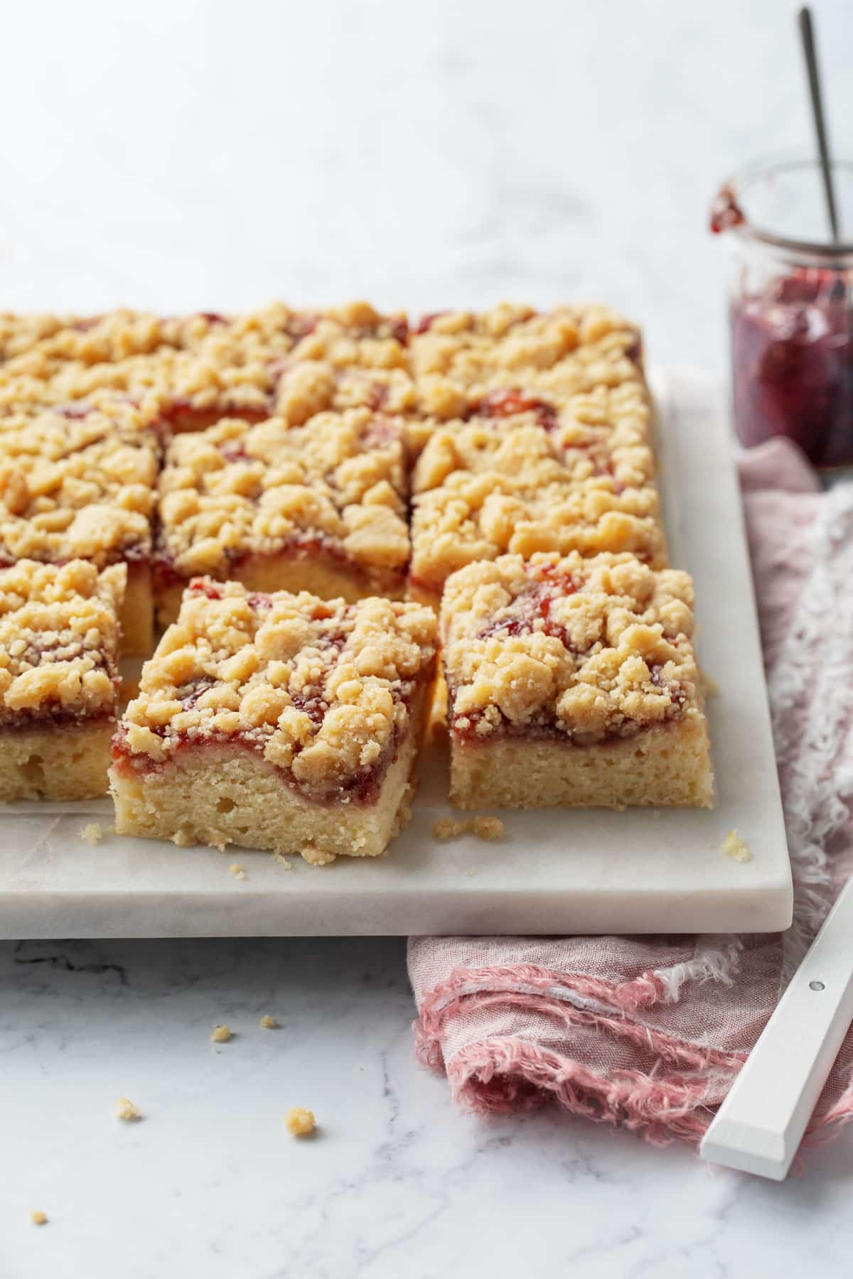 Jammy Sour Cream Crumb Cake cut into squares on a marble serving plate, with napkin, knife, and jar of jam on the side.