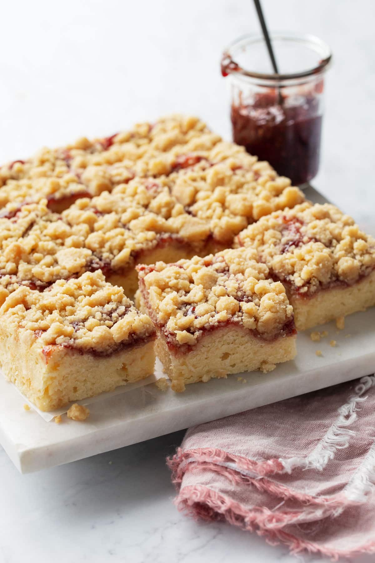 Jammy Sour Cream Crumb Cake cut into squares on a marble serving plate, with napkin, knife, and jar of jam on the side.