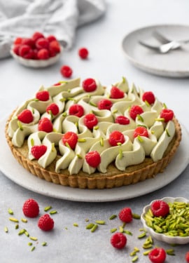 Raspberry & Pistachio Frangipane Tart with Whipped Pistachio Ganache piped in waves on a flat cake plate with rasberries and slivered pistachios scattered on top and around.