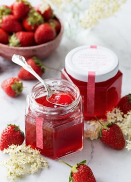 Two jars of Strawberry Elderflower Jelly, one open with a spoonful sitting on top to show the jelly texture, with fresh strawberries and elderflowers scattered around.