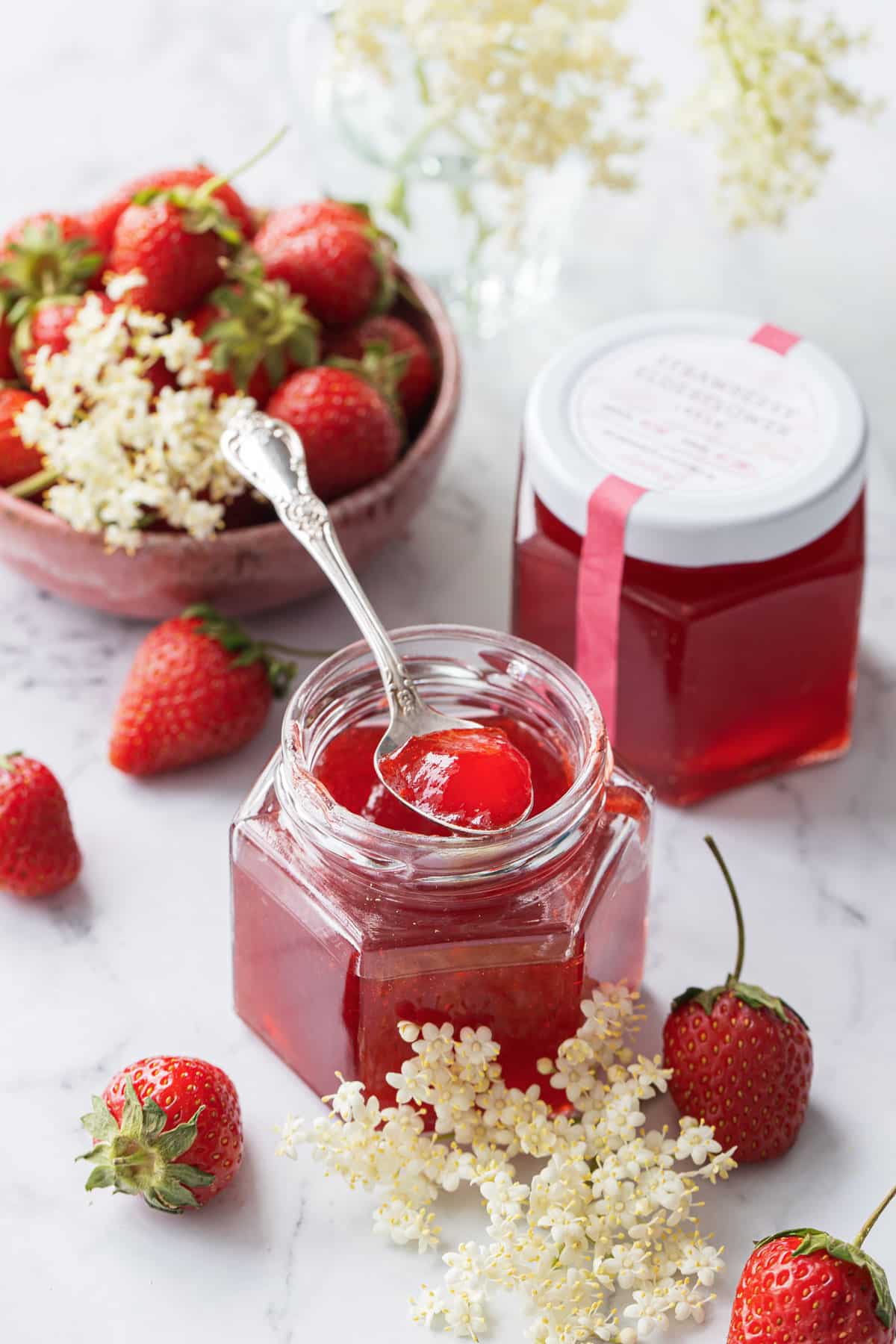 Two jars of Strawberry Elderflower Jelly, one open with a spoonful sitting on top to show the jelly texture, with a small bowl of strawberries in the background and sprigs of elderflowers scattered around.
