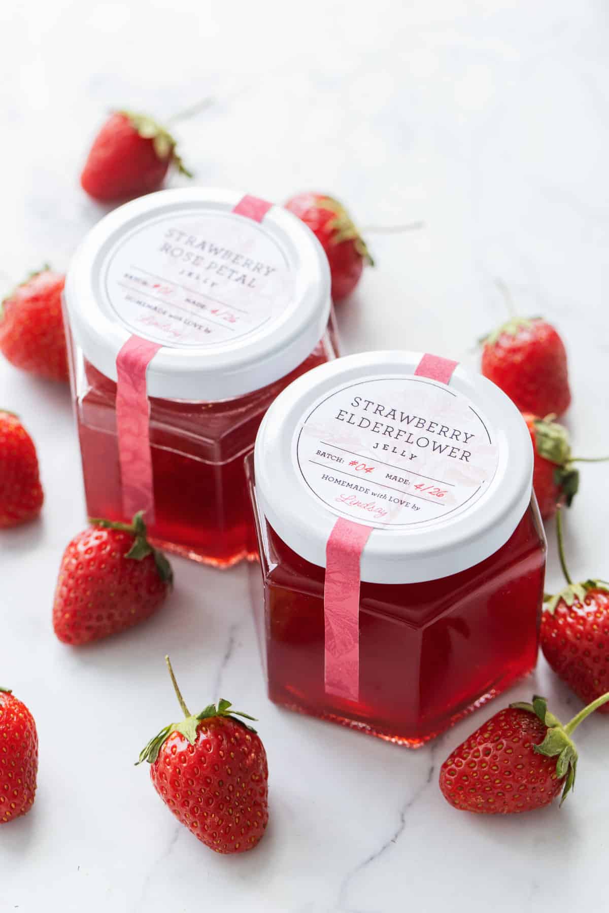 Two jars of Strawberry Elderflower and Strawberry Rose Jelly with custom printed labels on the lids.