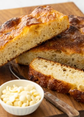 Slices of White Chocolate Focaccia Bread on a wooden cutting board with a bowl of white chocolate pieces on the side.