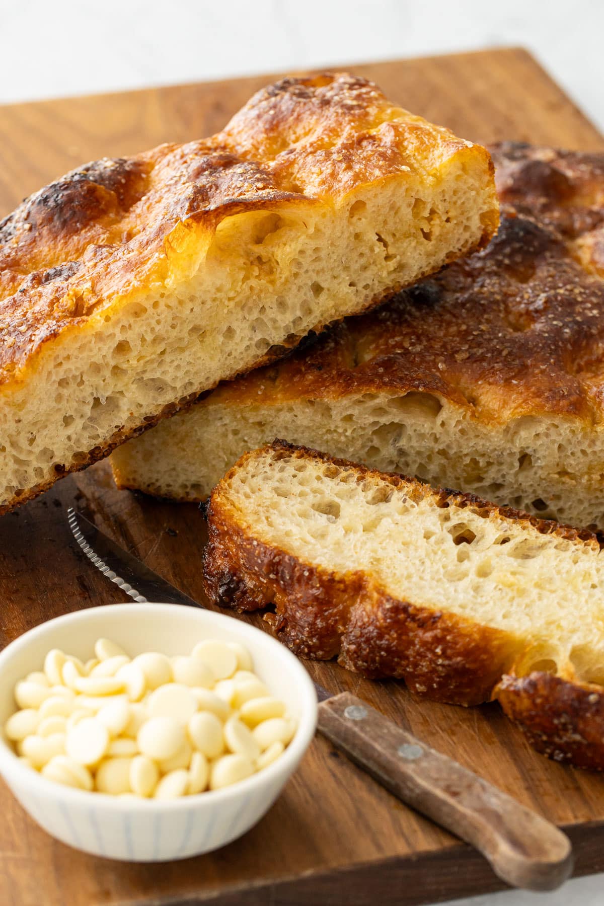 Slices of White Chocolate Focaccia Bread on a wooden cutting board with a bowl of white chocolate pieces on the side.