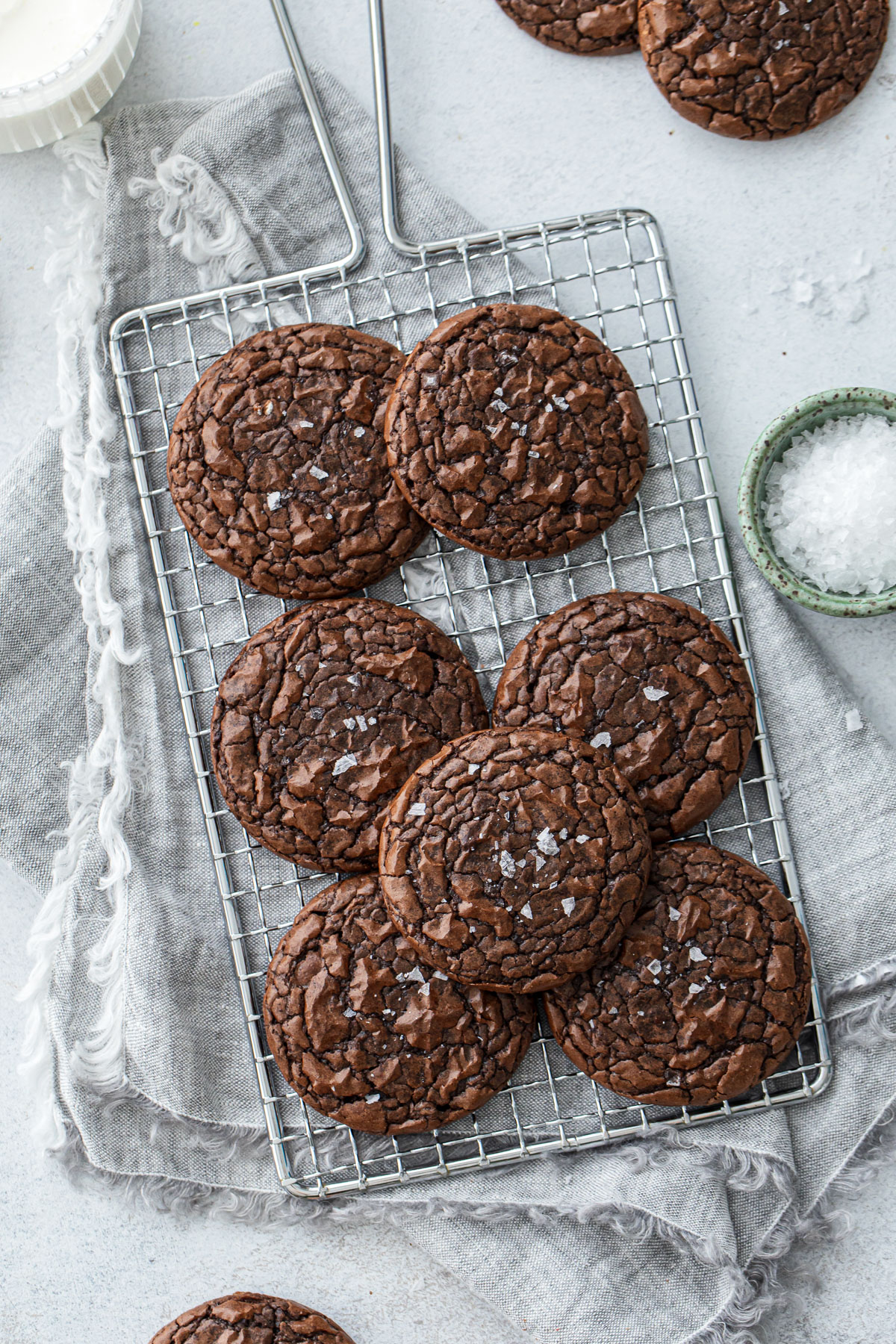 Chewy Crinkle-Top Brownie Cookies on a small wire baking rack, with linen napkin and a bowl of flaky sea salt on the side.