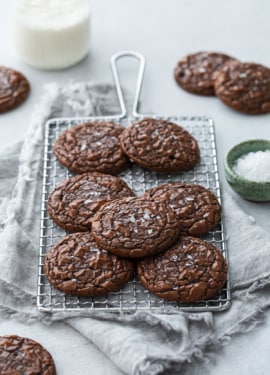Chewy Crinkle-Top Brownie Cookies on a small wire baking rack, with linen napkin and a bowl of flaky sea salt on the side.