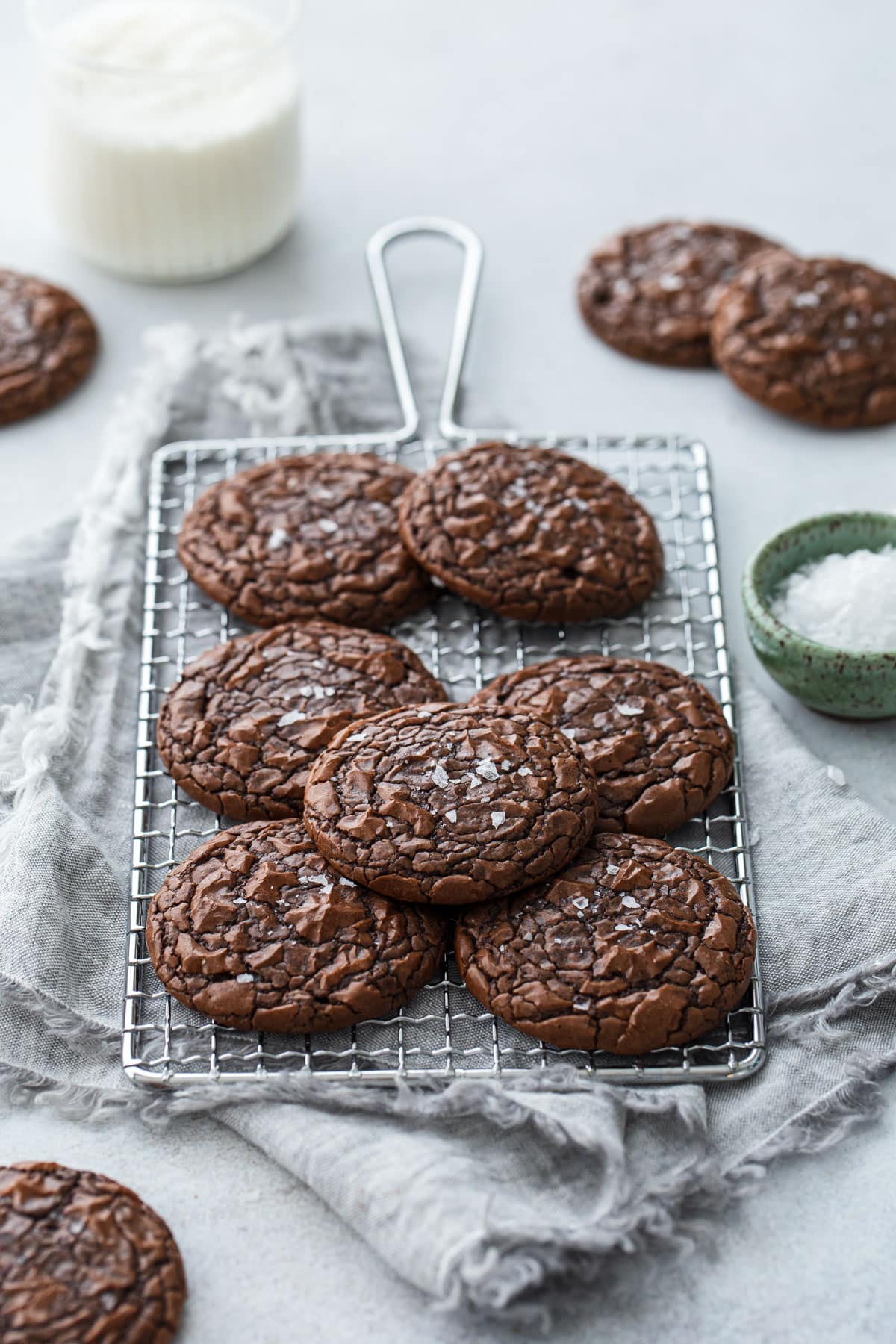 Chewy Crinkle-Top Brownie Cookies on a small wire baking rack, with linen napkin and a bowl of flaky sea salt on the side.