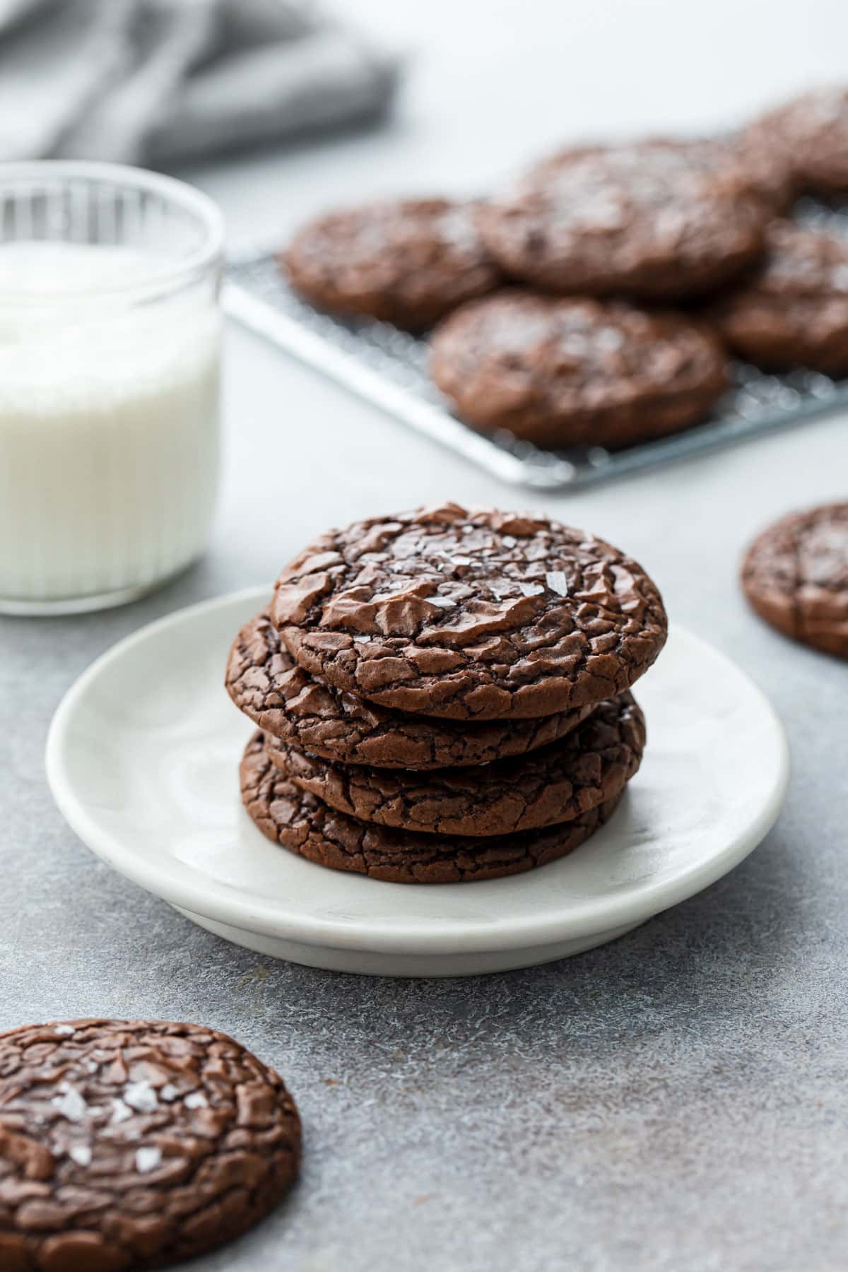Stack of Chewy Crinkle-Top Brownie Cookies on a plate with one cookie propped on the rim to show the shiny, crackly top texture.