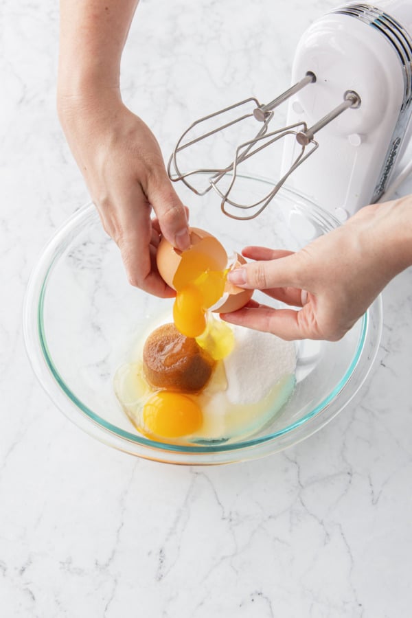 Cracking an egg into a glass mixing bowl with sugar and brown sugar in it.