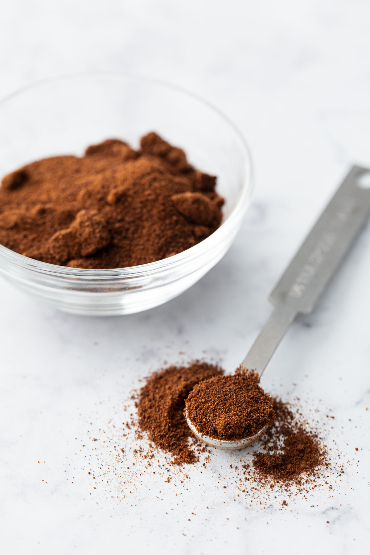 Clear bowl with instant espresso powder, with a quarter teaspoon full of more espresso powder on the side.