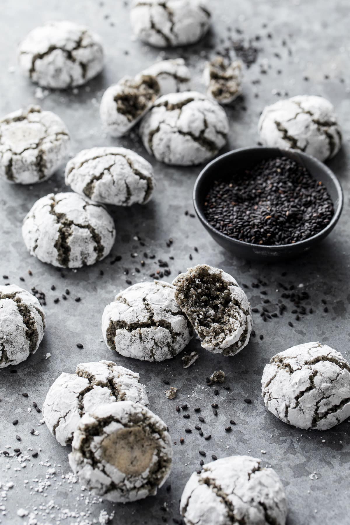 Black Sesame Amaretti Cookies scattered messily on a gray background with a bowl of black sesame seeds.