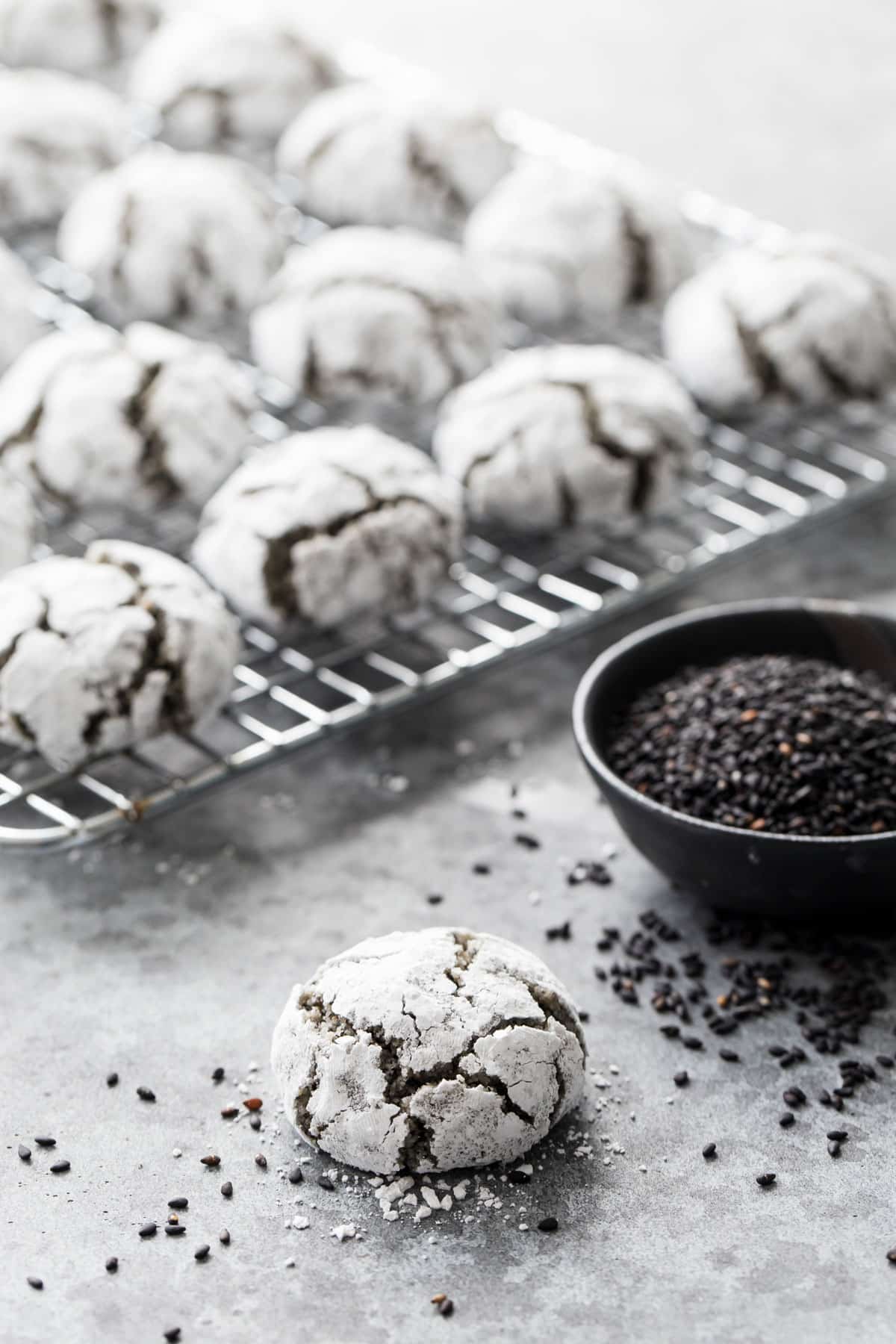 One Black Sesame Amaretti Cookie in the foreground showing the crackly, powdered-sugar coated appearance, with a bowl of black sesame seeds and a cooling rack with more cookies in the background.