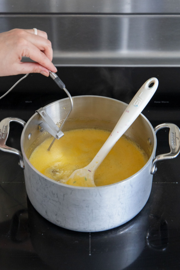 Placing a probe thermometer on the side of the saucepan filled with boiling passionfruit candy mixture.