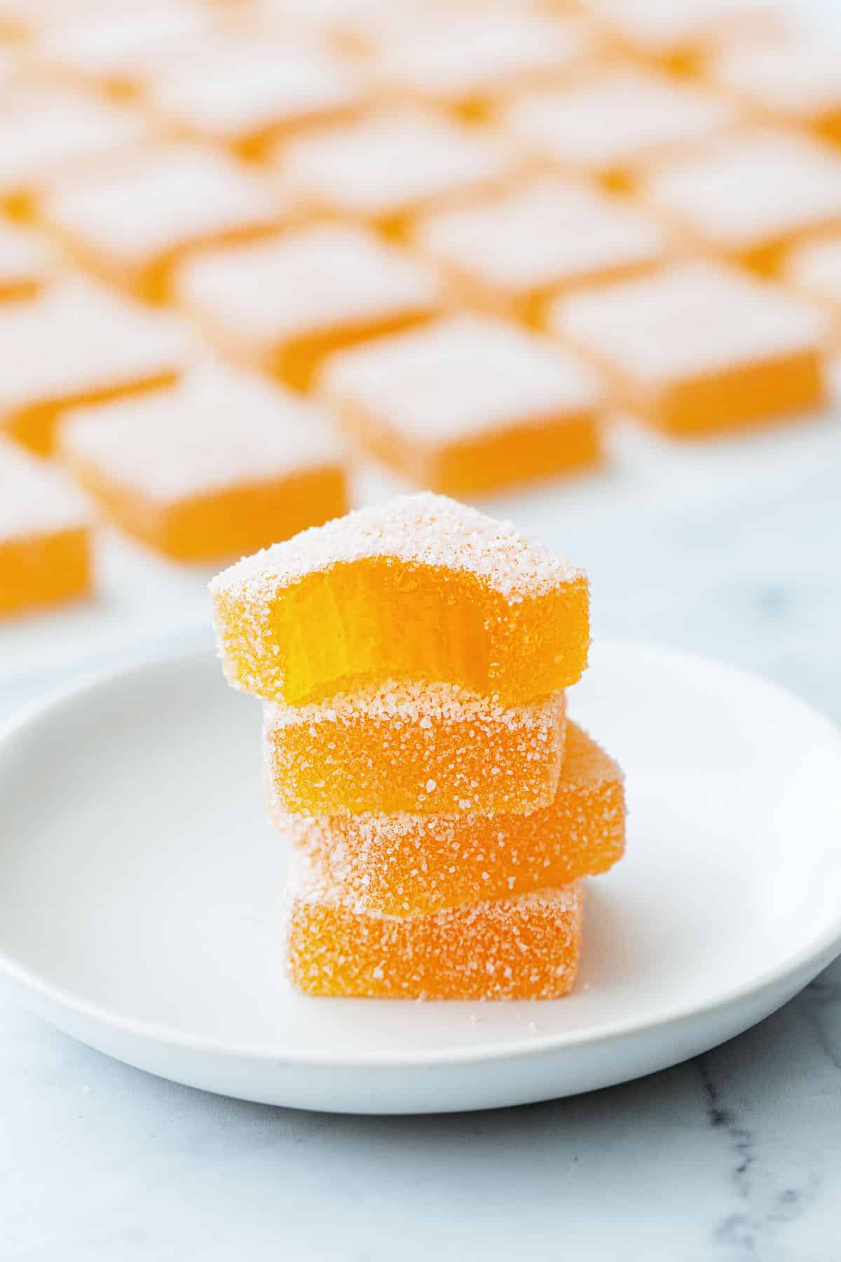Stack of four square pieces of Passionfruit Pate de Fruit on a small white plate, with more candies in the background.