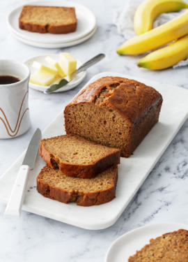 Loaf of Brown Butter Banana Bread with two slices laying down, on a marble platter with coffee, butter, and bananas completing the scene.