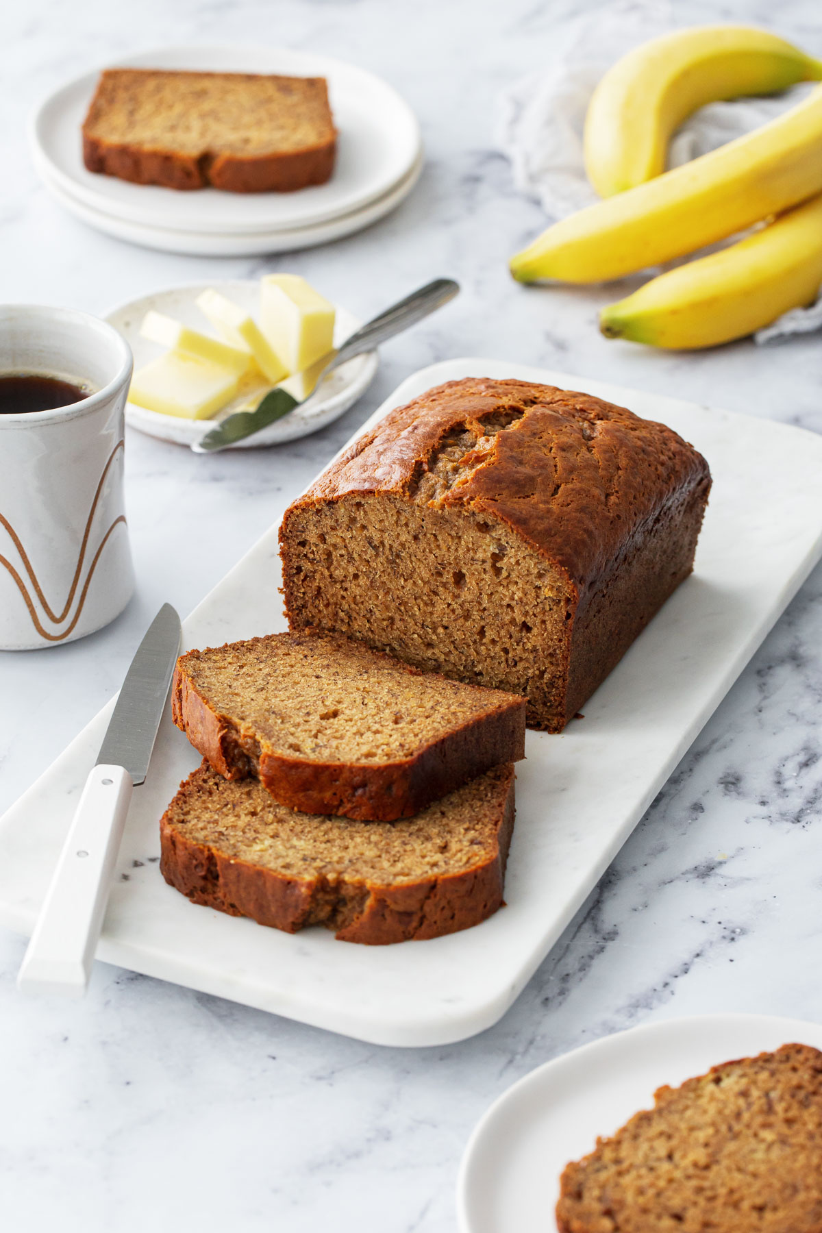 Loaf of Brown Butter Banana Bread with two slices laying down, on a marble platter with coffee, butter, and bananas completing the scene.