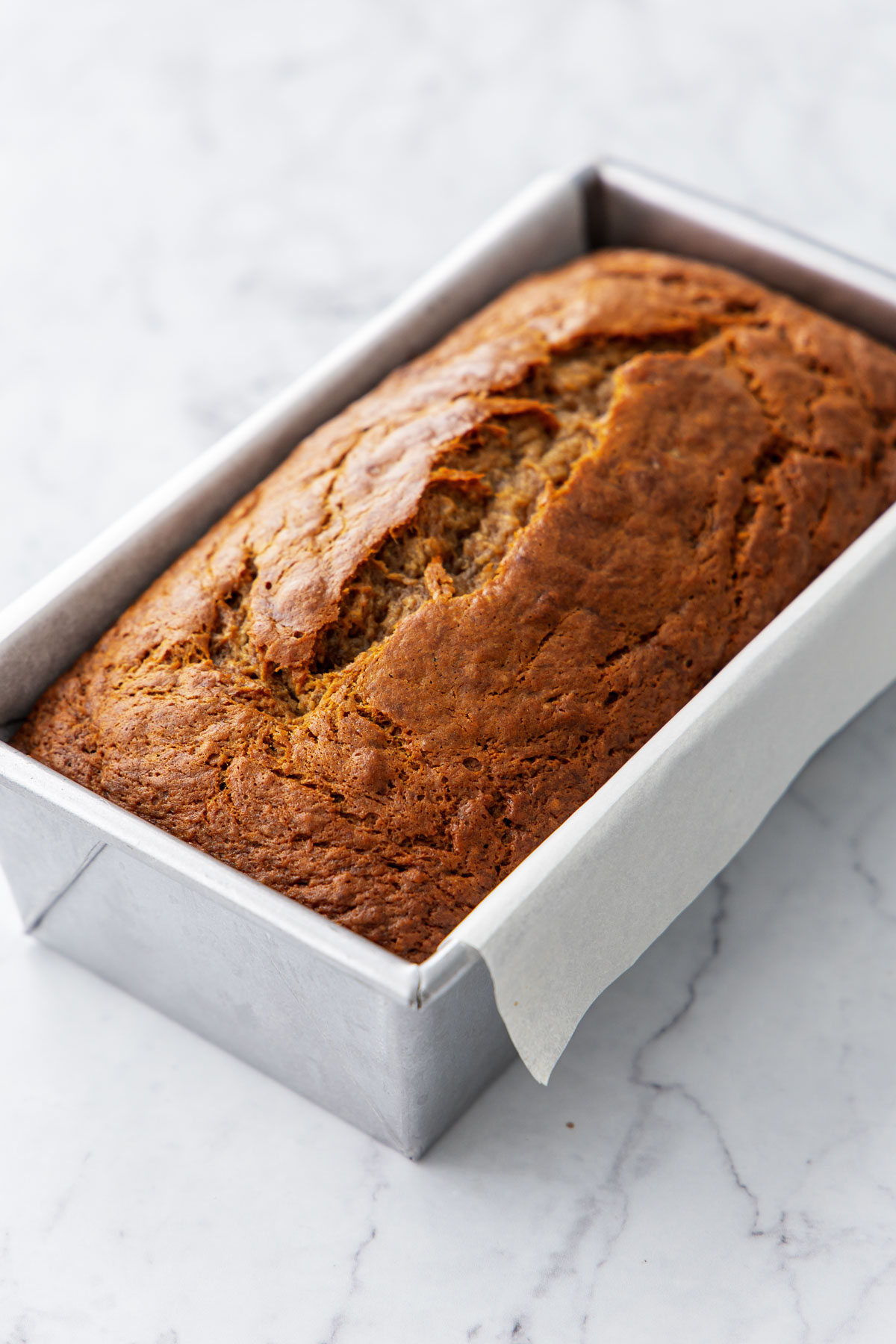 Loaf of Brown Butter Banana Bread in a light metal baking pan, with a prominent crack going down the middle and a golden brown crust.