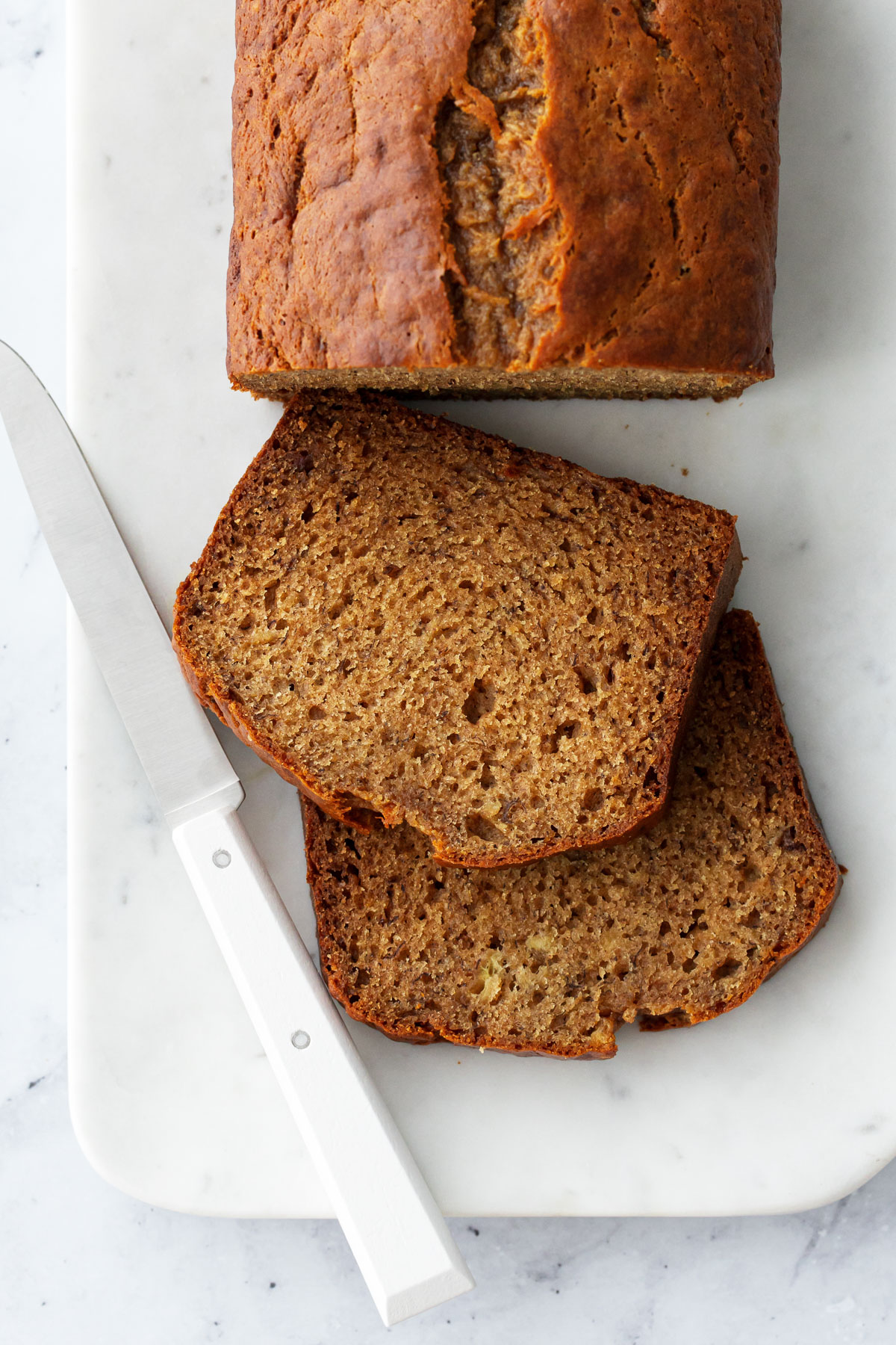 Loaf of Brown Butter Banana Bread on a marble platter, two cut slices laying down to show the interior texture, white knife on the side.