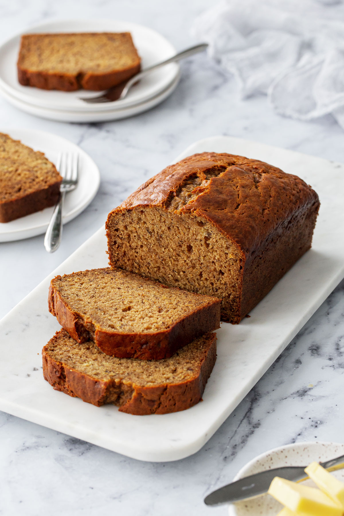 Loaf of Brown Butter Banana Bread with two slices laying down, on a marble platter with coffee, butter, and bananas completing the scene.
