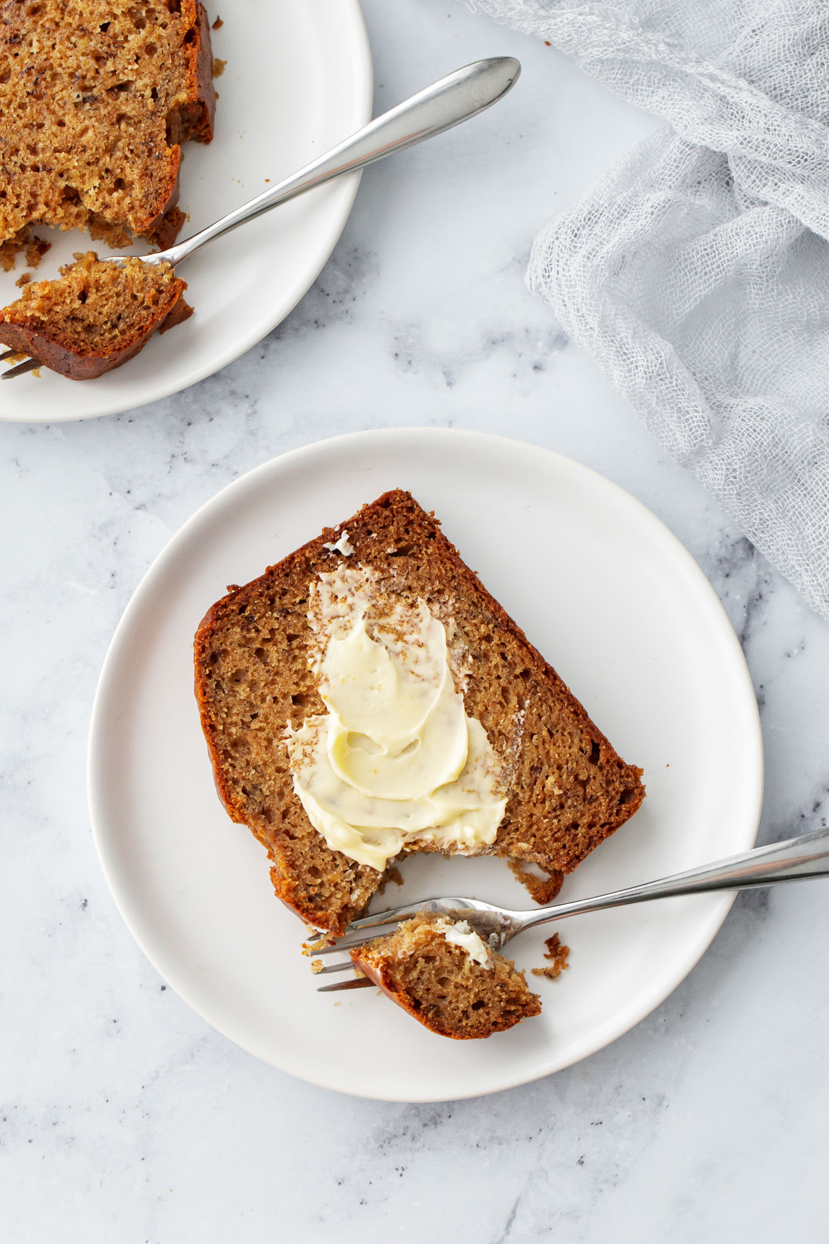 Overhead, slice of Brown Butter Banana Bread slathered with butter, fork cutting a bite to show the moist, rich texture of the bread.