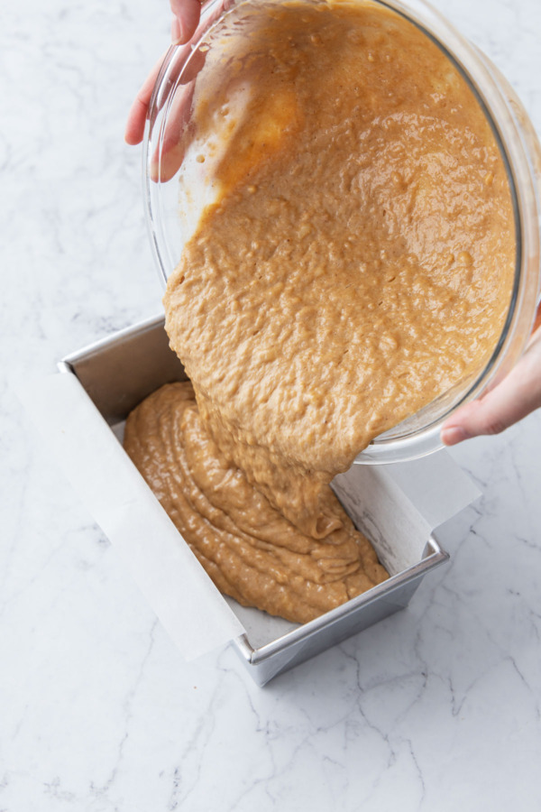 Pouring finished banana bread batter into parchment-lined metal loaf pan.