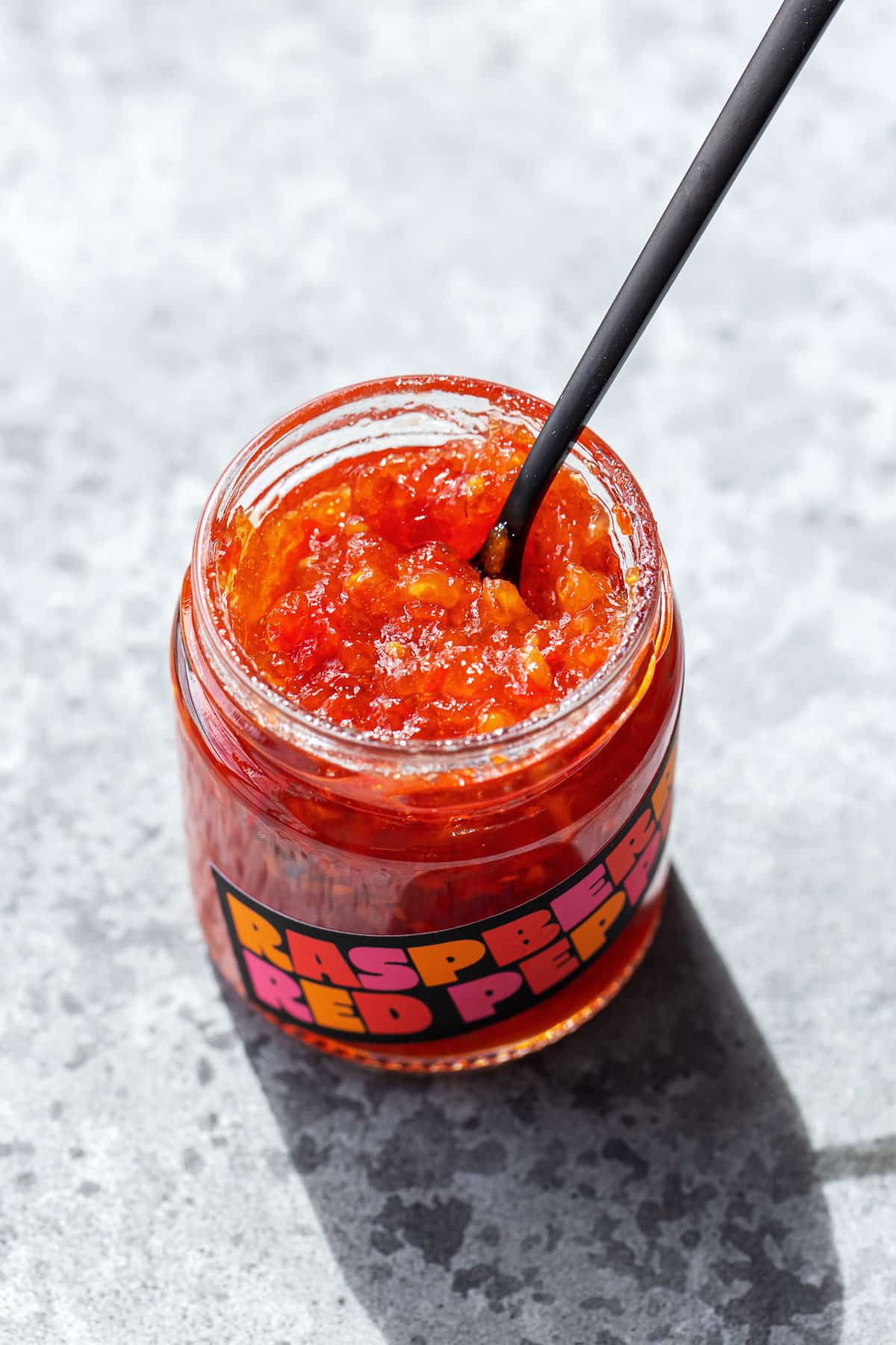 Glass jar of Raspberry Red Pepper Jelly, open with a spoon sticking out of it to show the texture of the bright red jelly with pieces of pepper visible.