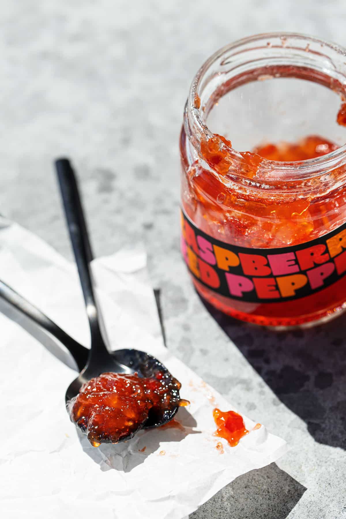 Two spoons with a dollop of Raspberry Red Pepper Jelly, showing the clarity and texture of the pepper-chunk-filled jelly, with an open jar on the side.