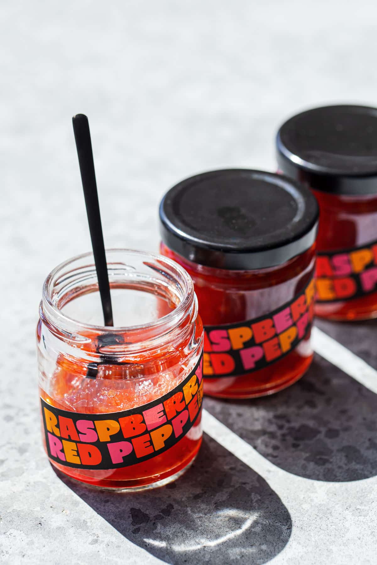 Three jars of Raspberry Red Pepper Jelly in a row, with harsh backlight creating strong shadows and backlighting the jelly to make it look like stained glass.