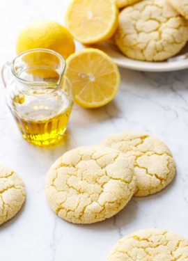 Crackly top Lemon Olive Oil Sugar Cookies on marble with a small glass pourer of olive oil, lemons, and a plate of more cookies in the background.