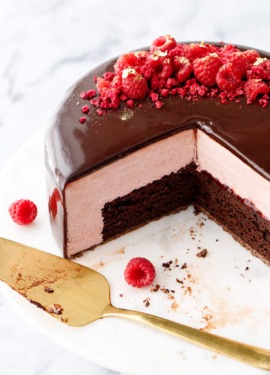 Raspberry and chocolate entremet-style cake with a dark chocolate mirror glaze with a few slices cut out to show the interior layers, with two raspberries and a gold cake server on the side.
