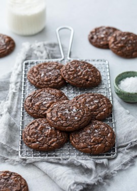 Chewy Crinkle-Top Brownie Cookies on a small wire baking rack, with linen napkin and a bowl of flaky sea salt on the side.
